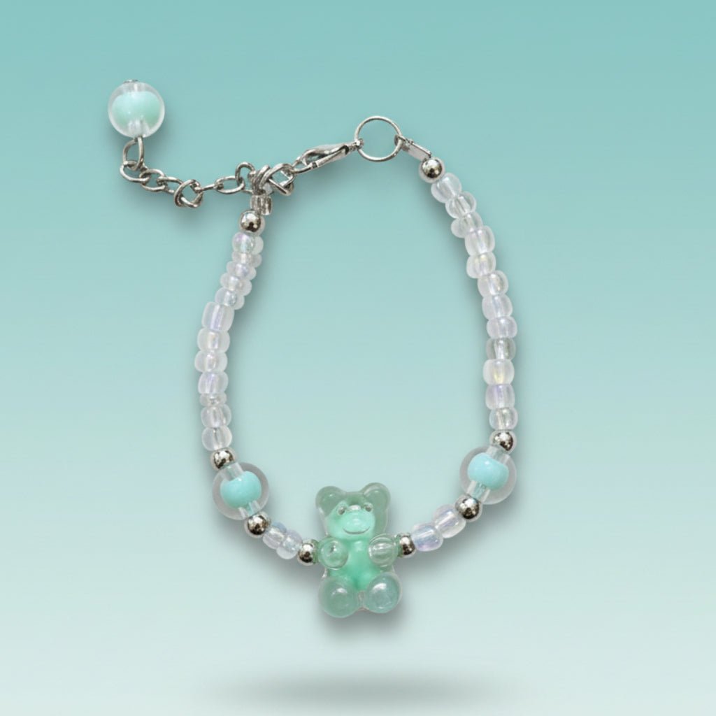 A handmade beaded charm bracelet featuring a translucent mint green resin teddy bear charm at the center. The bracelet is crafted with alternating iridescent clear beads, small silver spacer beads, and two large accent mint green round beads on either side. It features a silver lobster clasp closure with an adjustable chain and a single mint green bead dangling from the end, all set against a soft, gradient light teal background.