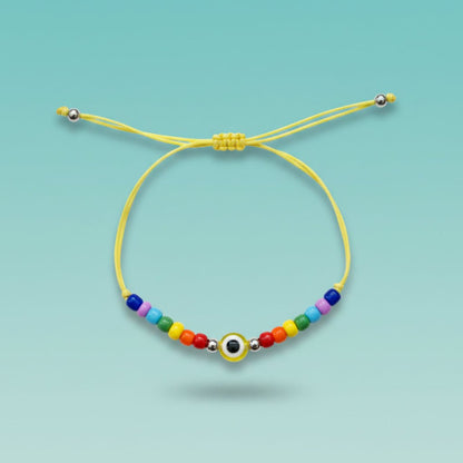 An adjustable yellow string bracelet featuring a central yellow and white evil eye charm, accented by small silver beads and a vibrant sequence of multi-colored beads on a light teal background.