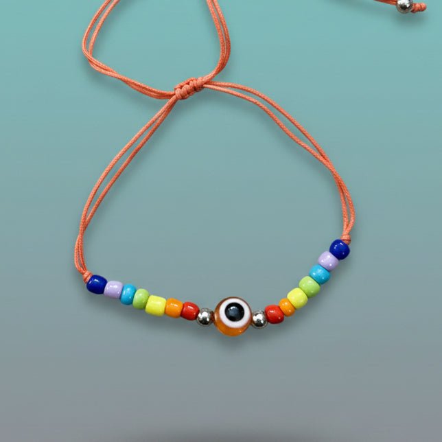 An adjustable orange string bracelet featuring a central orange and white evil eye charm, accented by small silver beads and a symmetrical sequence of rainbow-colored beads on a light teal background.