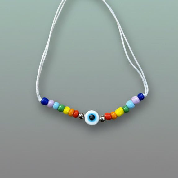 A colorful bead bracelet featuring a central white and blue evil eye charm, flanked by a rainbow arrangement of red, yellow, green, light blue, purple, and dark blue beads on a white cord.