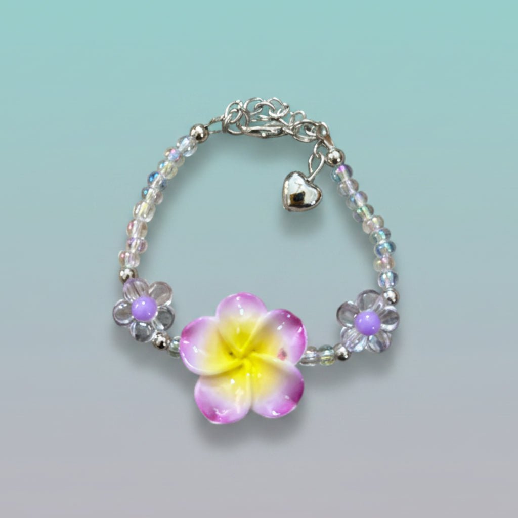 Nature-inspired beaded bracelet featuring a prominent purple and yellow plumeria flower centerpiece, clear iridescent beads, two small purple flower accents, and a silver heart charm on a light teal background.