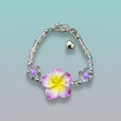 Nature-inspired beaded bracelet featuring a prominent purple and yellow plumeria flower centerpiece, clear iridescent beads, two small purple flower accents, and a silver heart charm on a light teal background.