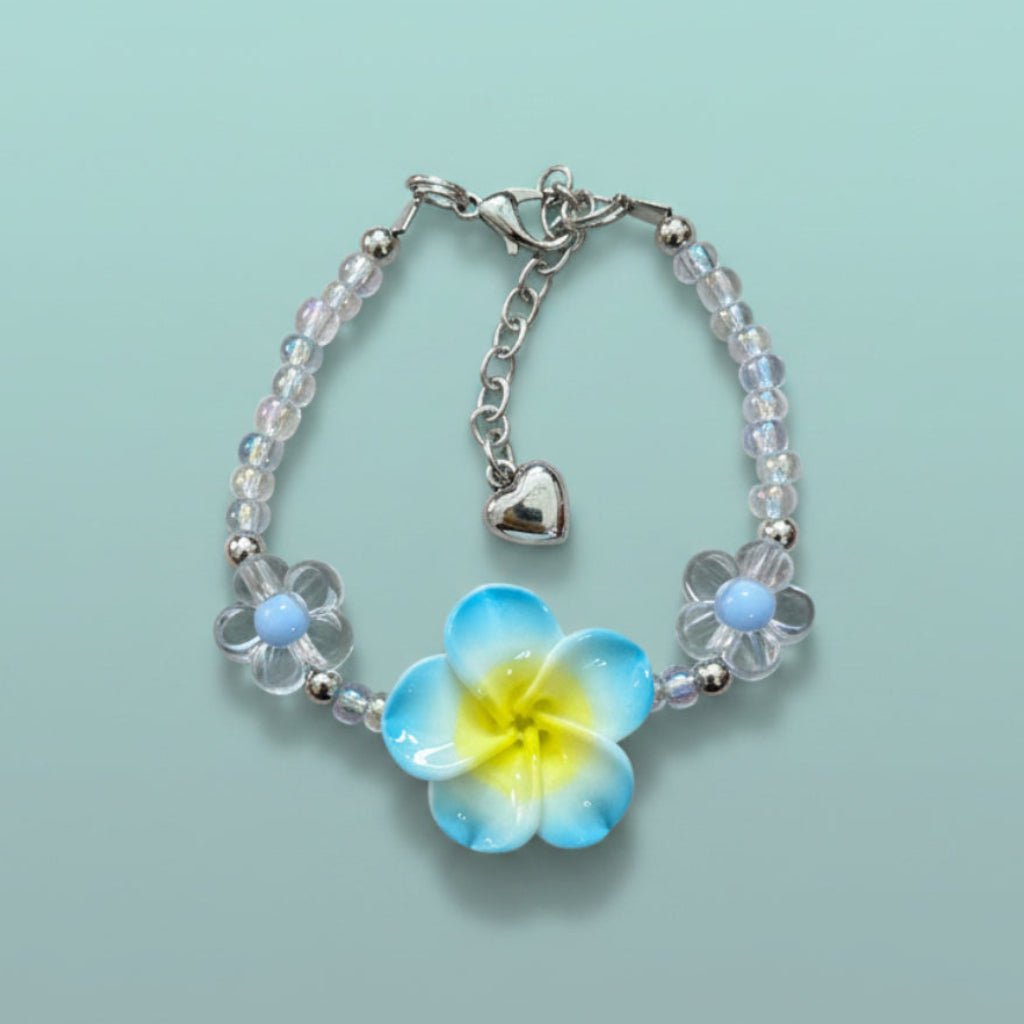 Delicate beaded floral charm bracelet with clear and light blue glass beads, a prominent yellow and blue flower charm, and a small silver heart pendant on a light blue background.