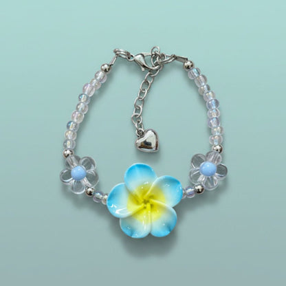 Delicate beaded floral charm bracelet with clear and light blue glass beads, a prominent yellow and blue flower charm, and a small silver heart pendant on a light blue background.