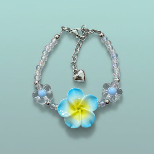 Delicate beaded floral charm bracelet with clear and light blue glass beads, a prominent yellow and blue flower charm, and a small silver heart pendant on a light blue background.