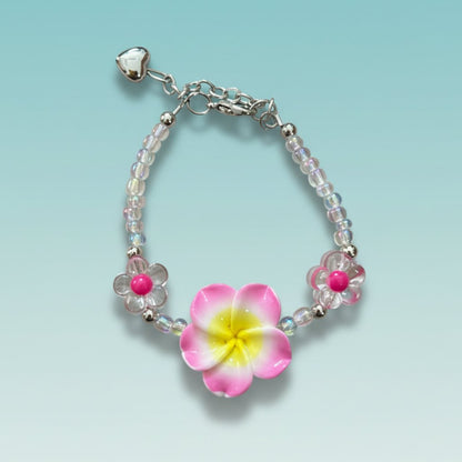 Beaded chain bracelet featuring a prominent pink and yellow tropical plumeria flower charm, clear iridescent beads, two small pink flower accents, and a silver heart charm on a light teal background.
