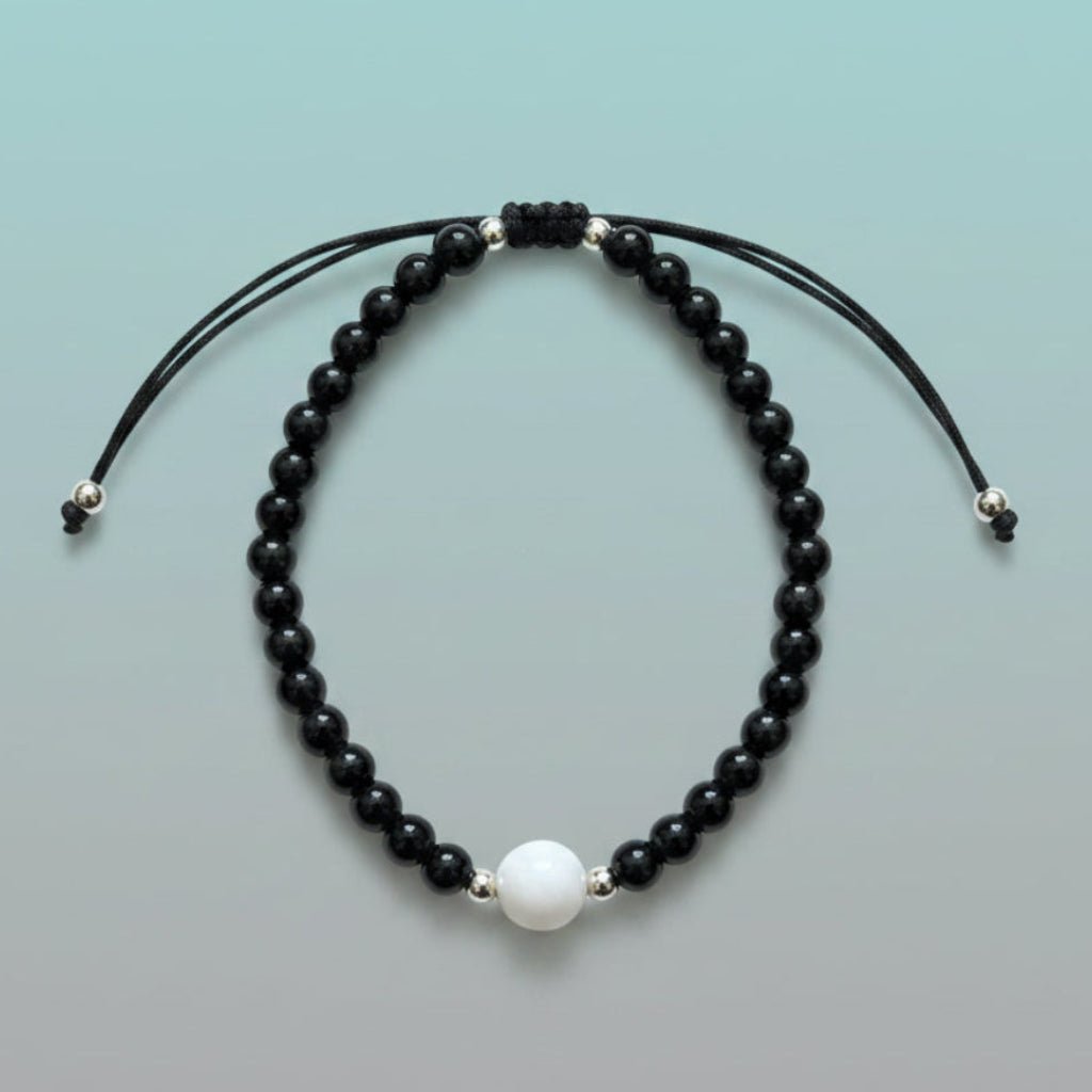 Handmade adjustable black string bracelet featuring a large central white pearl, accented by a strand of glossy black beads and silver spacers on a soft mint green background.