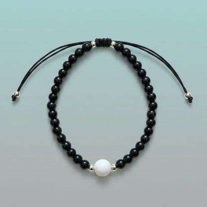 Handmade adjustable black string bracelet featuring a large central white pearl, accented by a strand of glossy black beads and silver spacers on a soft mint green background.