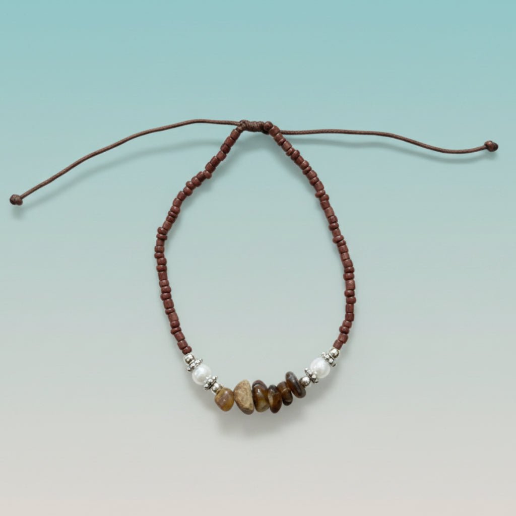 Brown cord seashell bracelet featuring reddish-brown seed beads, white pearls, and earthy brown stone accents on a light blue background.
