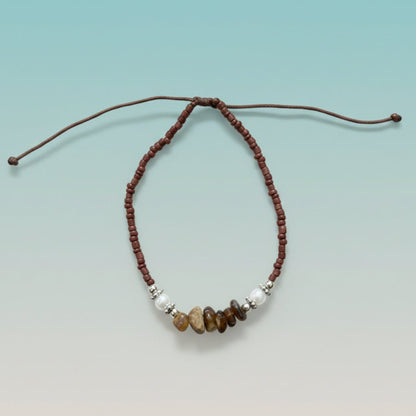Brown cord seashell bracelet featuring reddish-brown seed beads, white pearls, and earthy brown stone accents on a light blue background.