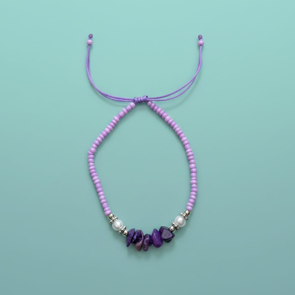 Purple cord bracelet featuring light purple seed beads, white pearls, and dark purple stone accents on a light blue background.