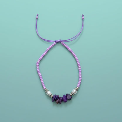 Purple cord bracelet featuring light purple seed beads, white pearls, and dark purple stone accents on a light blue background.