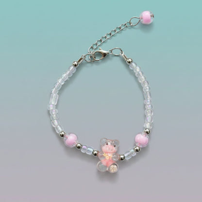A handmade beaded charm bracelet featuring a small, translucent pink and orange resin teddy bear pendant at the center. The bracelet is crafted with clear, silver, and pastel pink round beads, and includes an adjustable silver chain clasp with a single pink bead dangling at the end.