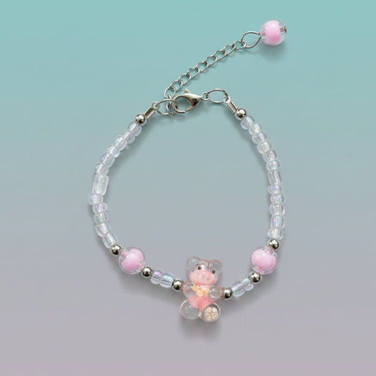 A handmade beaded charm bracelet featuring a small, translucent pink and orange resin teddy bear pendant at the center. The bracelet is crafted with clear, silver, and pastel pink round beads, and includes an adjustable silver chain clasp with a single pink bead dangling at the end.