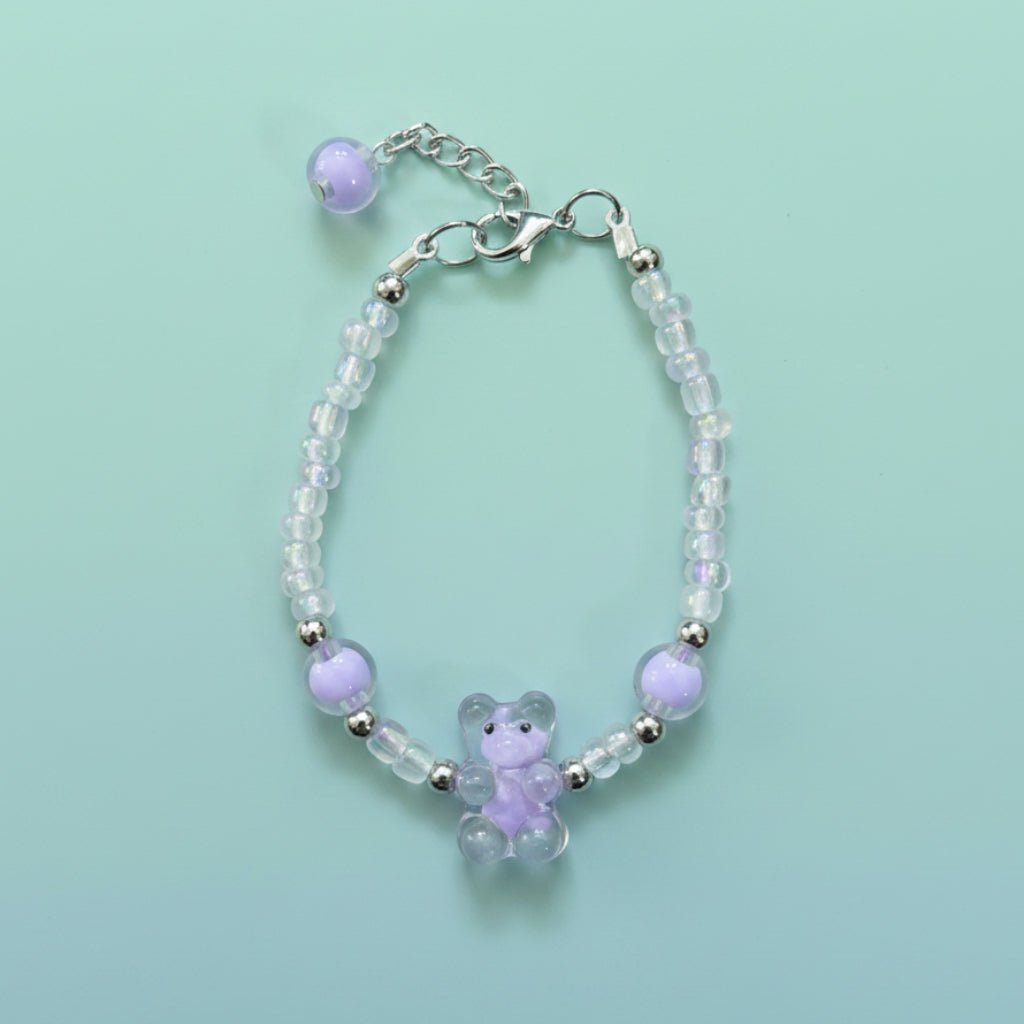 A handmade beaded charm bracelet featuring a translucent lavender purple resin teddy bear charm at the center. The bracelet is crafted with alternating iridescent clear beads, small silver spacer beads, and two large accent purple round beads on either side. It features a silver lobster clasp closure with an adjustable chain and a single purple bead dangling from the end, all set against a soft, solid mint green background.