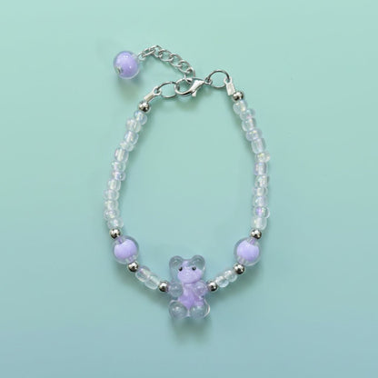 A handmade beaded charm bracelet featuring a translucent lavender purple resin teddy bear charm at the center. The bracelet is crafted with alternating iridescent clear beads, small silver spacer beads, and two large accent purple round beads on either side. It features a silver lobster clasp closure with an adjustable chain and a single purple bead dangling from the end, all set against a soft, solid mint green background.