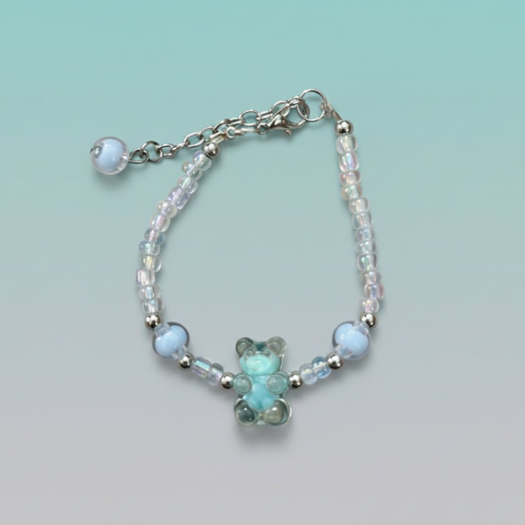A handmade beaded charm bracelet featuring a translucent light blue resin teddy bear charm at the center. The bracelet is crafted with alternating iridescent clear beads, small silver spacer beads, and two large accent light blue round beads on either side. It features a silver lobster clasp closure with an adjustable chain and a single light blue bead dangling from the end, all set against a soft, gradient mint green background.