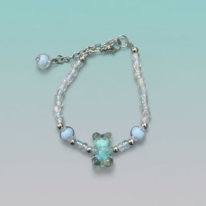 A handmade beaded charm bracelet featuring a translucent light blue resin teddy bear charm at the center. The bracelet is crafted with alternating iridescent clear beads, small silver spacer beads, and two large accent light blue round beads on either side. It features a silver lobster clasp closure with an adjustable chain and a single light blue bead dangling from the end, all set against a soft, gradient mint green background.