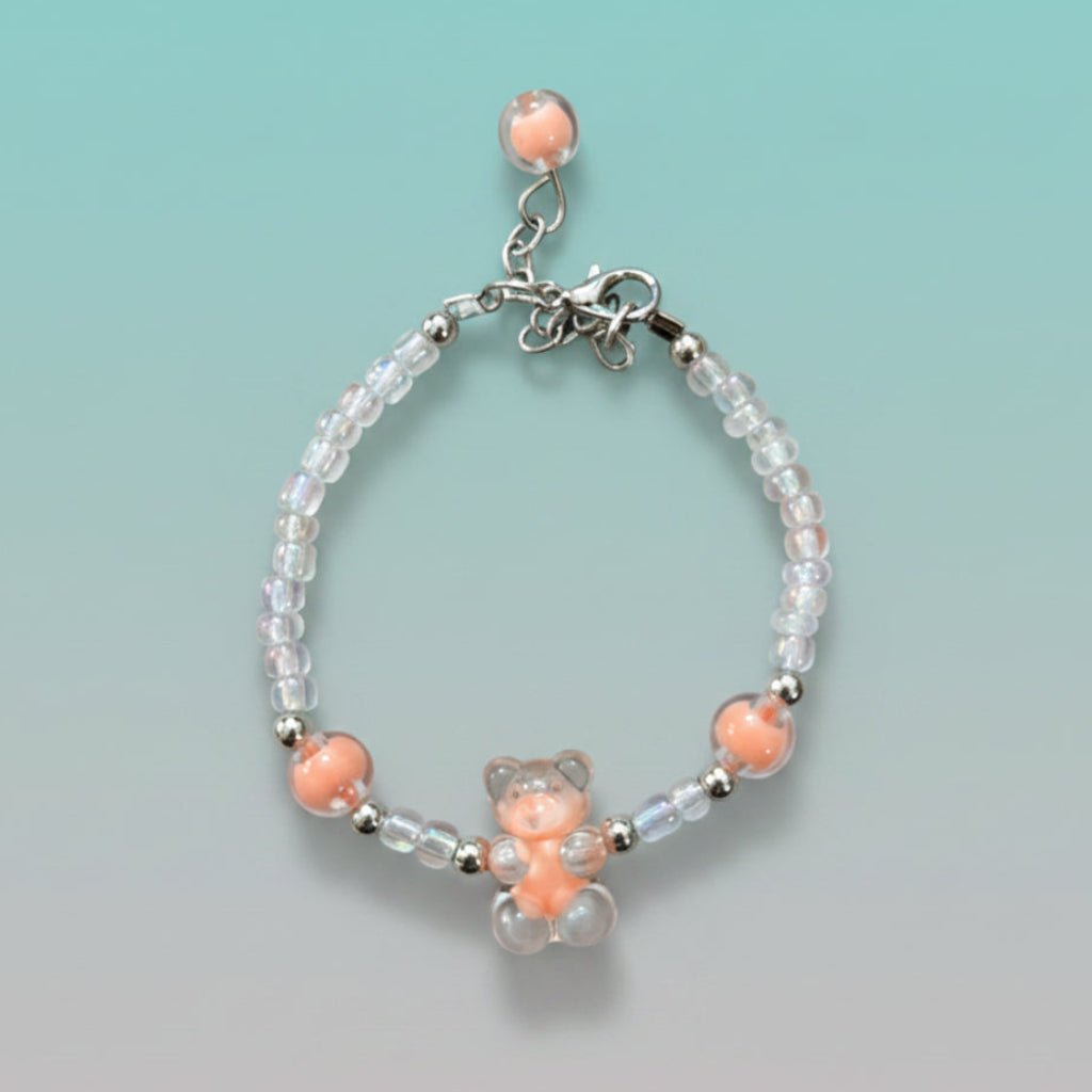 A handmade beaded charm bracelet featuring a translucent pastel peach resin teddy bear charm at the center. The bracelet is crafted with alternating iridescent clear beads, small silver spacer beads, and two large accent peach round beads on either side. It features a silver lobster clasp closure with an adjustable chain and a single peach bead dangling from the end, all set against a soft, gradient mint green background.