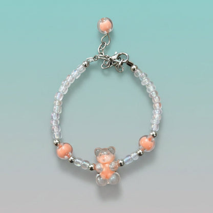 A handmade beaded charm bracelet featuring a translucent pastel peach resin teddy bear charm at the center. The bracelet is crafted with alternating iridescent clear beads, small silver spacer beads, and two large accent peach round beads on either side. It features a silver lobster clasp closure with an adjustable chain and a single peach bead dangling from the end, all set against a soft, gradient mint green background.