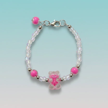 A handmade beaded charm bracelet featuring a translucent hot pink resin teddy bear charm at the center. The bracelet is crafted with alternating iridescent clear beads, small silver spacer beads, and two large accent hot pink round beads on either side. It features a silver lobster clasp closure with an adjustable chain and a single hot pink bead dangling from the end, all set against a soft, gradient mint green background.