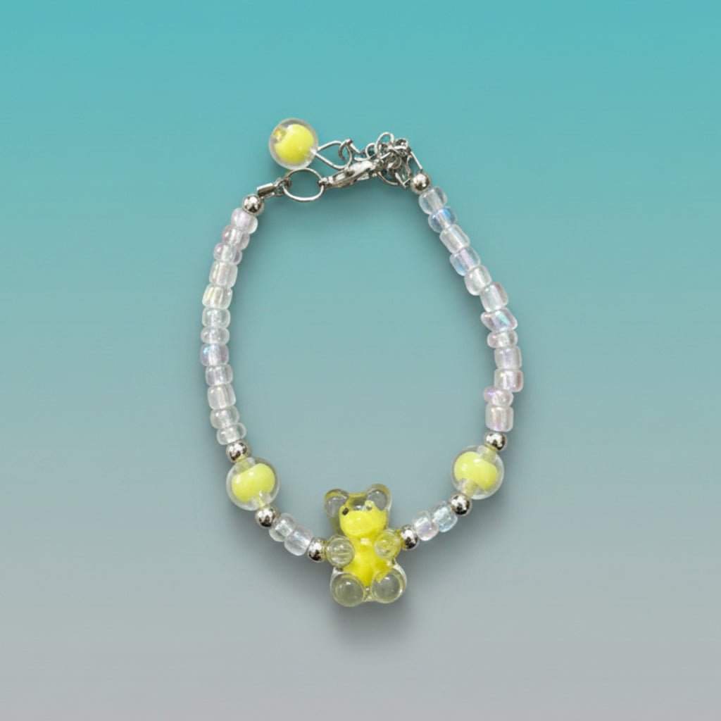 A handmade beaded charm bracelet featuring a translucent bright yellow resin teddy bear charm at the center. The bracelet is crafted with alternating iridescent clear beads, small silver spacer beads, and two large accent yellow round beads on either side. It features a silver lobster clasp closure with an adjustable chain and a single yellow bead dangling from the end, all set against a soft, gradient teal and mint green background.