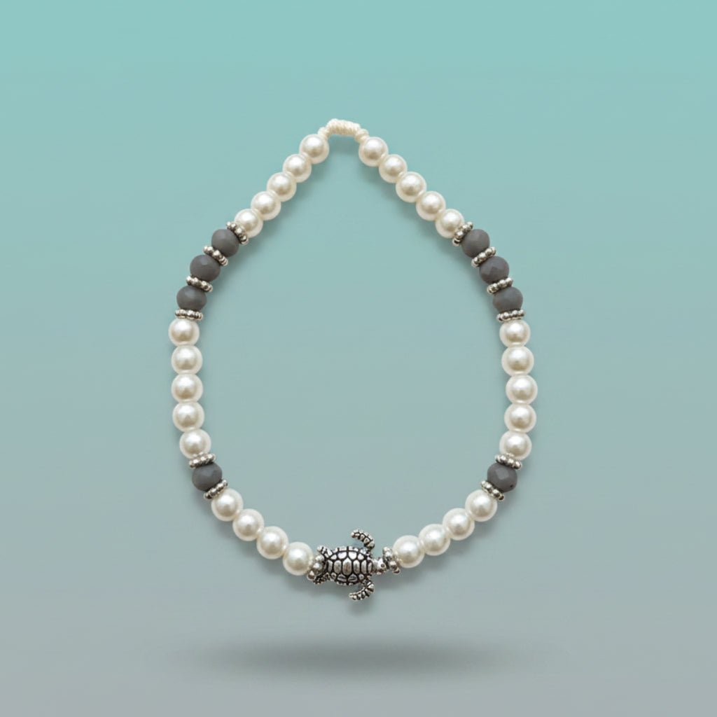 Elegantly patterned turtle bracelet featuring iridescent white pearl beads accented with textured dark-grey disc beads and a central silver turtle charm, set against a soft light-blue background.