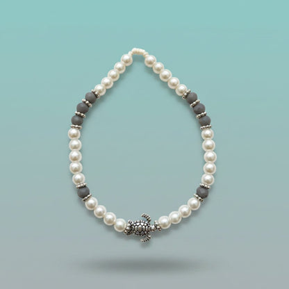 Elegantly patterned turtle bracelet featuring iridescent white pearl beads accented with textured dark-grey disc beads and a central silver turtle charm, set against a soft light-blue background.